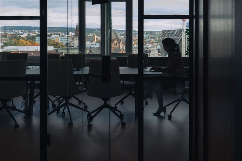 Person sitter og jobber ved en laptop i et møterom med glassvegger, med utsikt over byen i bakgrunnen.