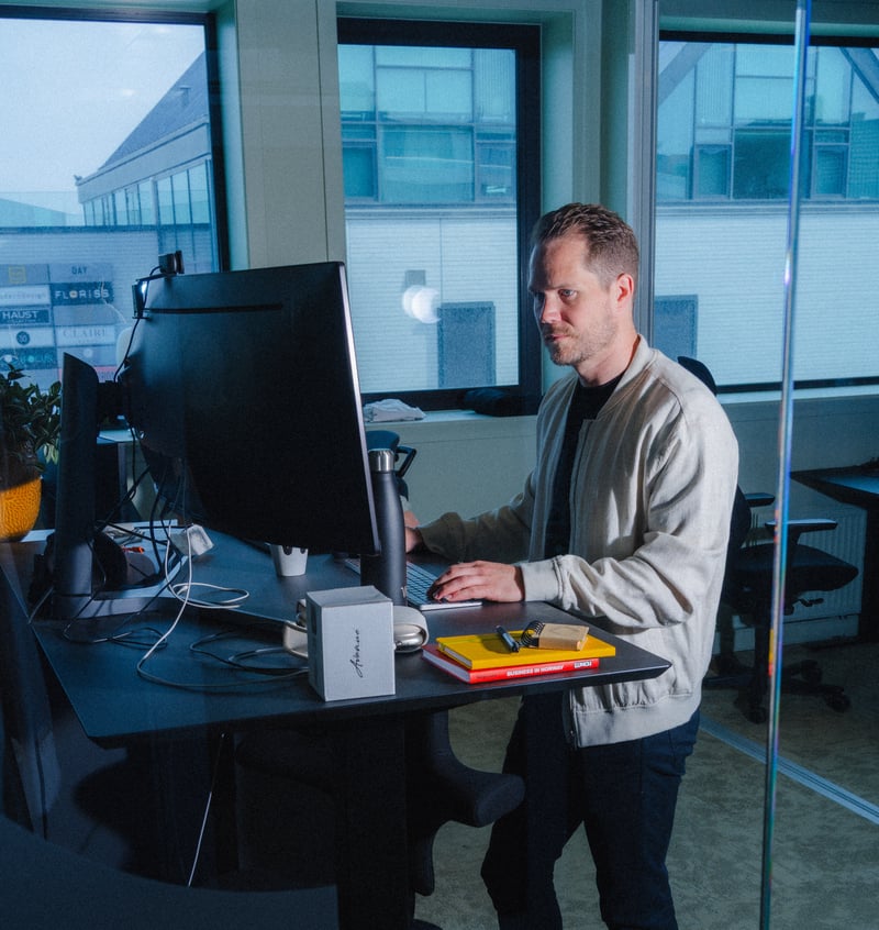 En person står ved et hev-senk-skrivebord og jobber konsentrert foran en stor skjerm i et kontormiljø.