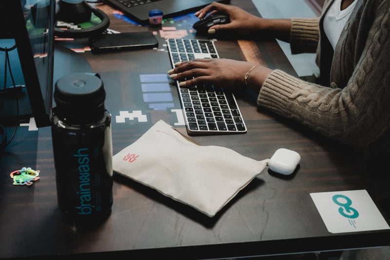 Hender på tastatur og datamus ved en arbeidsstasjon i kontormiljø, med skjerm, notater og arbeidsutstyr på skrivebordet.